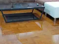 Wasserpfütze auf dem Holzboden, der Parkettboden ist aufgequollen und beschädigt. Ein Couchtisch steht im nassen Bereich.