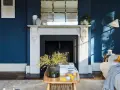 Ein Wohnzimmer mit tiefblauen Wänden. Ein Marmorkamin mit Spiegel darüber. Ein Holztisch mit Blumen und Spielzeug. Passt beige zu blau? Ja, die gelben Akzente und der beigefarbene Teppich zeigen es.