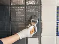 Hand mit Handschuh streicht mit Pinsel graue Farbe auf Fliesen. Ein Teil der Wand ist bereits grau, der andere noch weiß.
