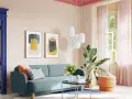 Ein Wohnzimmer mit hellrosa Decke und Wand, blauem Sofa, bunten Bildern und einem Durchgang zu einem Schlafzimmer.
