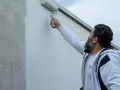 Ein Mann mit Bart streicht mit einer Malerrolle eine Hauswand. Die Wand zeigt horizontale Streifen, da sie gerade neu gestrichen wird.