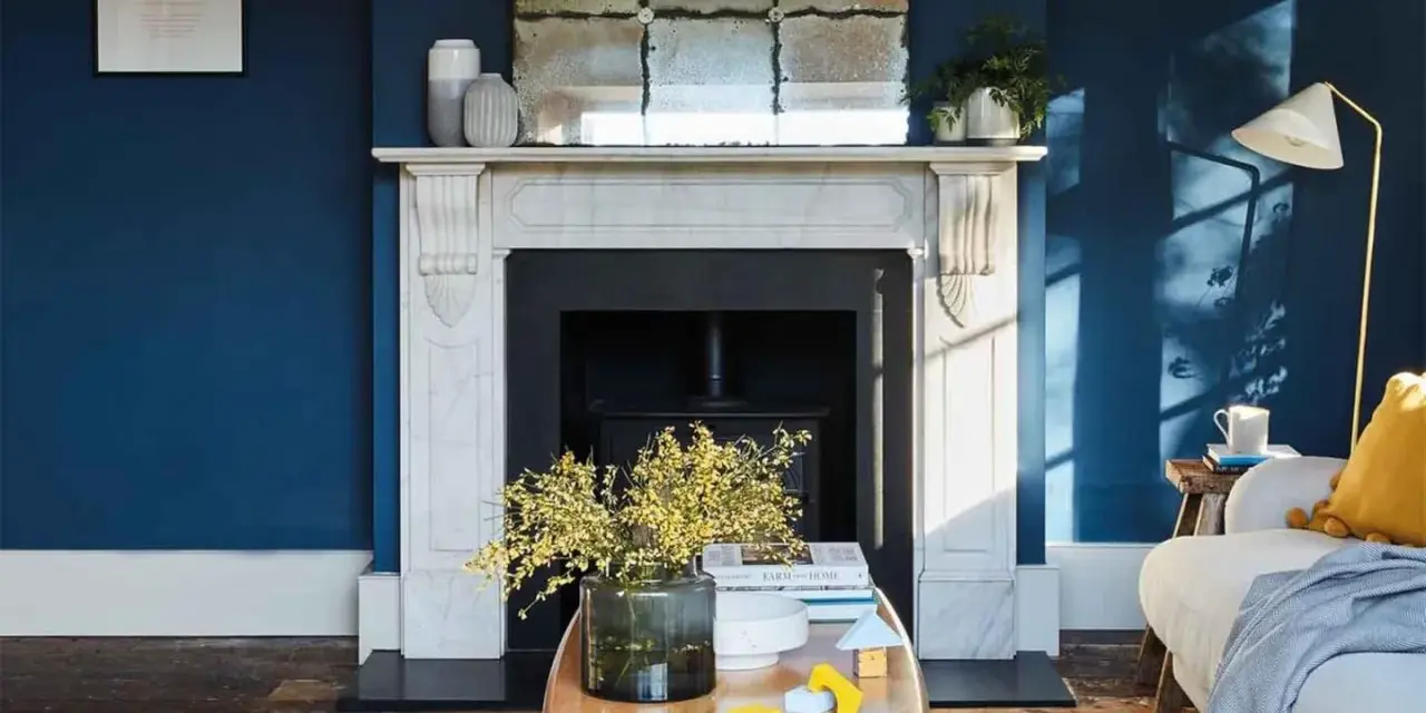 Ein Wohnzimmer mit tiefblauen Wänden. Ein Marmorkamin mit Spiegel darüber. Ein Holztisch mit Blumen und Spielzeug. Passt beige zu blau? Ja, die gelben Akzente und der beigefarbene Teppich zeigen es.
