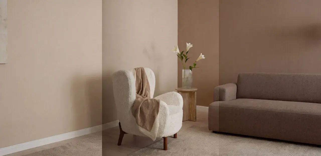 Gemütliches Wohnzimmer mit einem weißen Teddy-Sessel, einer braunen Couch und einer Vase mit Lilien. Warme Farben dominieren die Szene.
