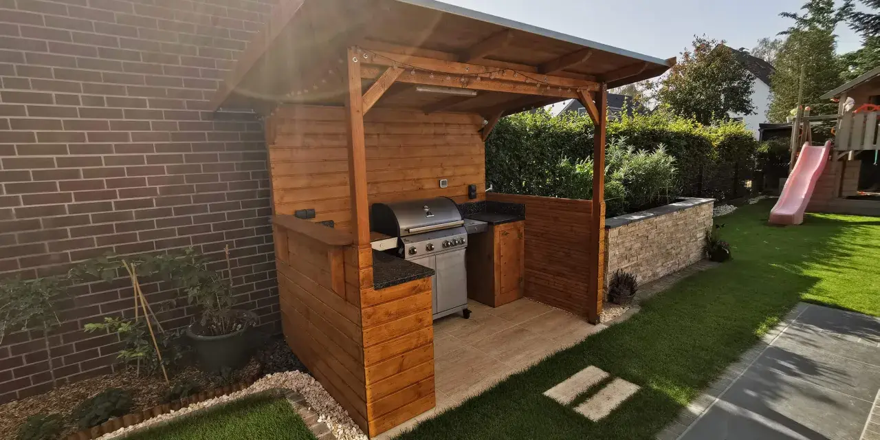Ein moderner Grillplatz im Garten mit überdachter Holzkonstruktion, Gasgrill und gemütlicher Beleuchtung. Perfekt für Sommerabende.