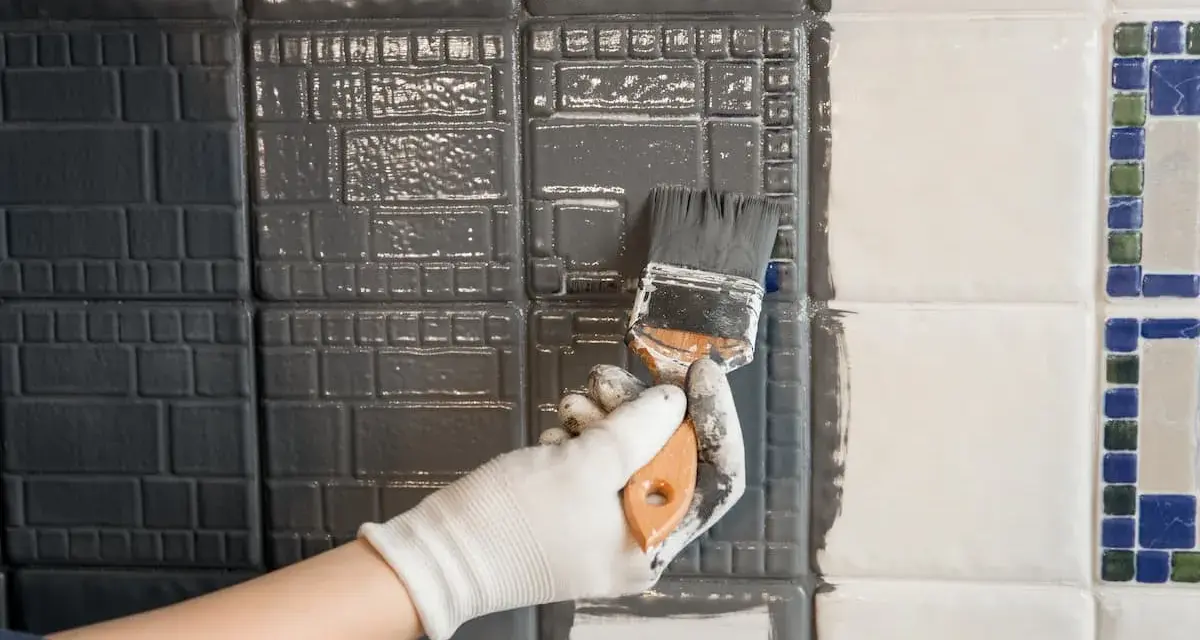Hand mit Handschuh streicht mit Pinsel graue Farbe auf Fliesen. Ein Teil der Wand ist bereits grau, der andere noch weiß.