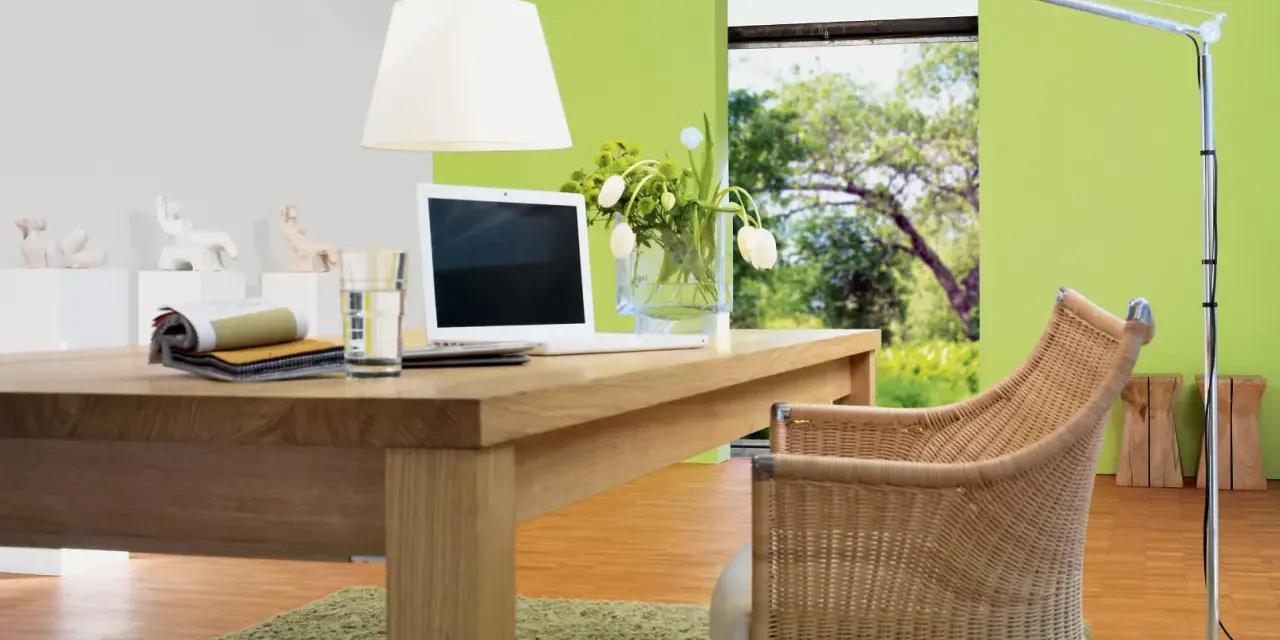 Ein Schreibtisch aus Nussbaumholz mit Laptop und Stoffmustern. Grüne Wand und Rattanstuhl. Welche Farbe passt zu Nussbaum? Grün und Beige.