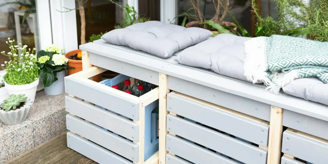 Ordnung im Garten: Eine graue Gartenbank mit Stauraum für Getränkekisten und Kissen. Pflanzen und eine Decke sorgen für Gemütlichkeit.
