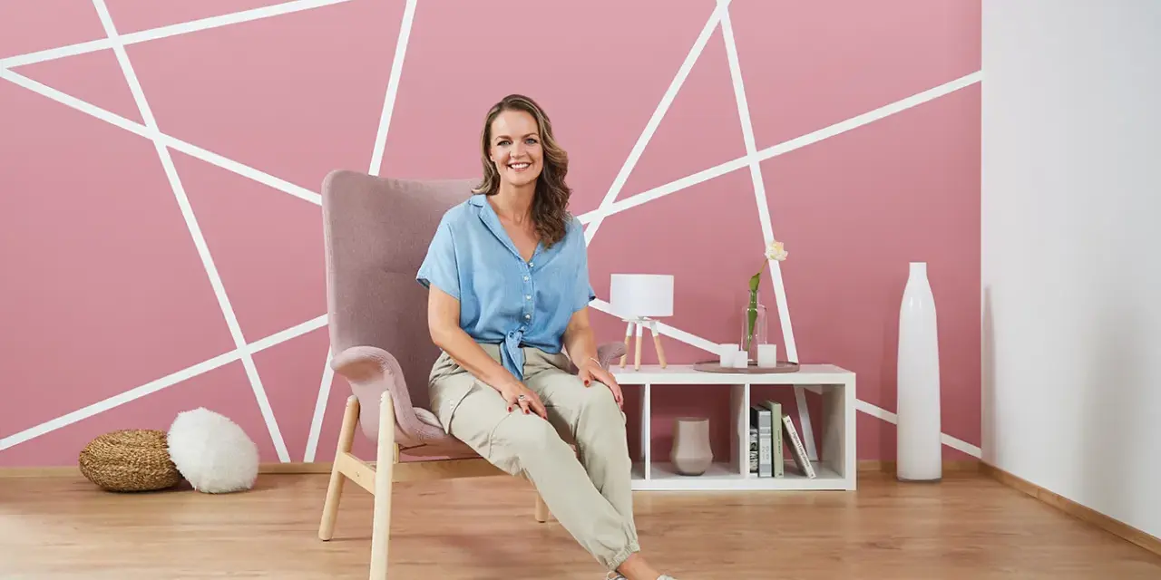 Frau sitzt vor einer rosa Wand mit weißem geometrischem Muster. Tolle Ideen zum Wand streichen und Muster abkleben!