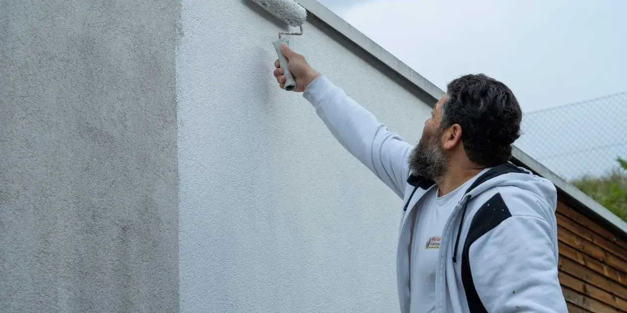 Ein Mann mit Bart streicht mit einer Malerrolle eine Hauswand. Die Wand zeigt horizontale Streifen, da sie gerade neu gestrichen wird.
