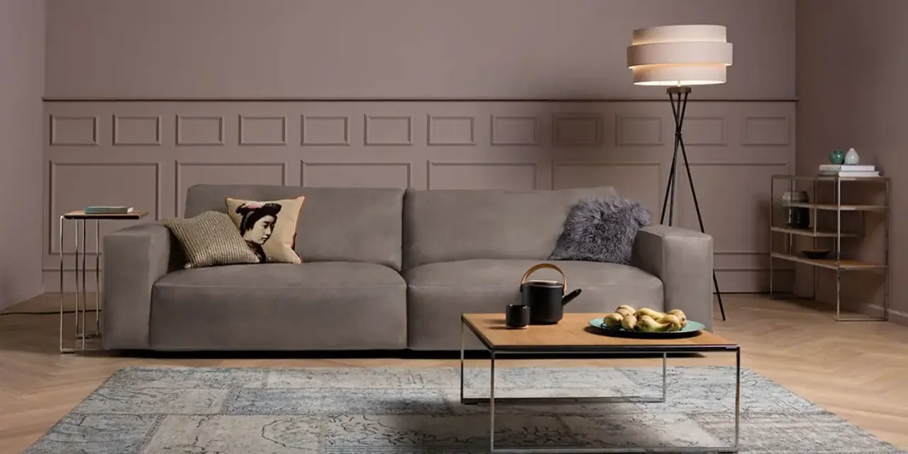 Ein taupefarbenes Sofa mit Kissen und ein Couchtisch mit Obst. Die Wandfarbe ist ein warmes Braun, das zeigt, welche Farbe passt zu Taupe.