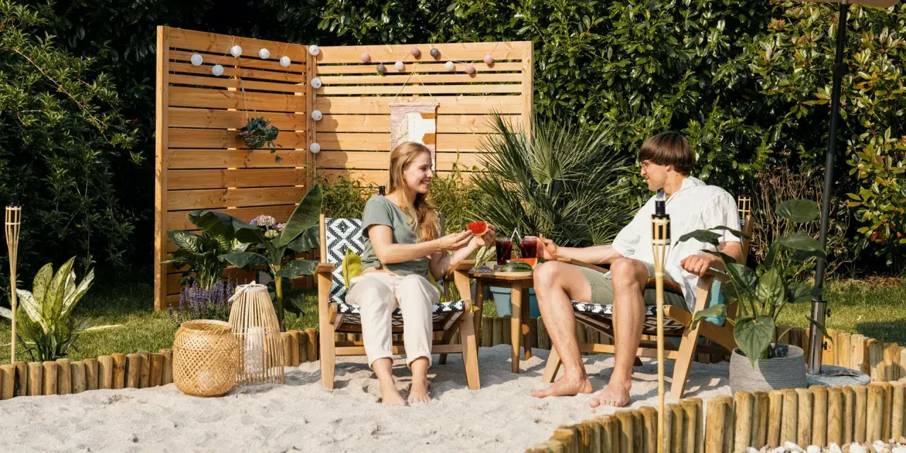 Ein Paar genießt den Sommer mit Wassermelone und Drinks im Garten. Sand, Holz und Pflanzen schaffen ein echtes Strandfeeling im Garten.