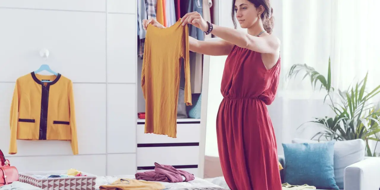 Frau sortiert Kleidung nach der Marie Kondo Methode. Ein gelbes Oberteil wird in die Hand genommen, daneben ein gelber Blazer und ein Korb mit gefalteten Textilien.