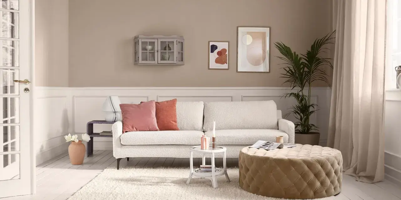 Gemütliches Wohnzimmer mit cremefarbener Couch, passenden Kissen und einem Hocker. Die ecru farbe kombinieren sich harmonisch mit den Wandfarben und Deko-Elementen.