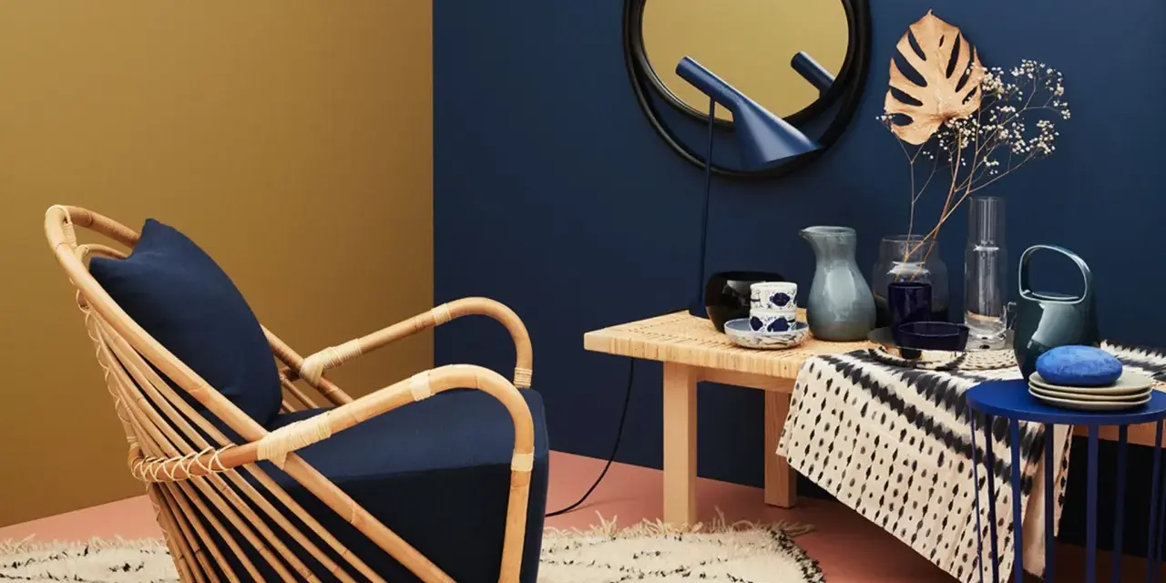 Ein Rattansessel mit dunkelblauem Kissen steht auf einem Teppich. Welche Farbe passt zu Gold? Das goldene Spiegelbild an der Wand und die gelbe Wandfarbe harmonieren mit dem tiefen Blau.