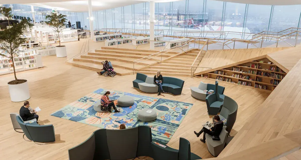 Helle, offene Bibliothek in Oodi Helsinki mit Menschen, die lesen und sich entspannen. Holzmöbel und große Fenster schaffen eine einladende Atmosphäre.
