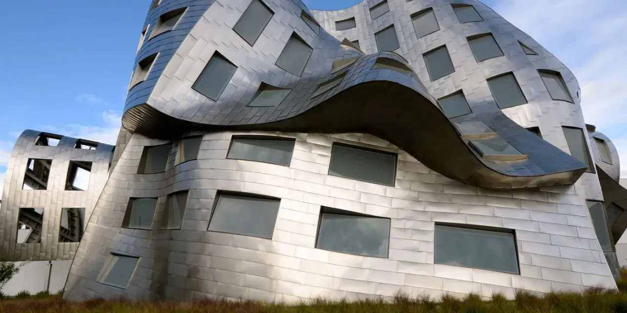 Ein futuristisches Gebäude mit geschwungenen Metallfassaden, typisch für Frank Gehry Bauwerke, ragt aus hohem Gras empor.