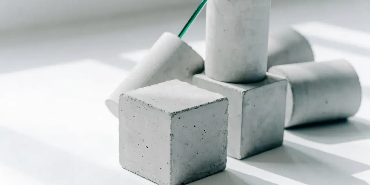 Betonwürfel und -zylinder in verschiedenen Formen, die die Vielfalt der **Beton festigkeitsklassen** symbolisieren. Ein grüner Stab ragt aus einem der Objekte.