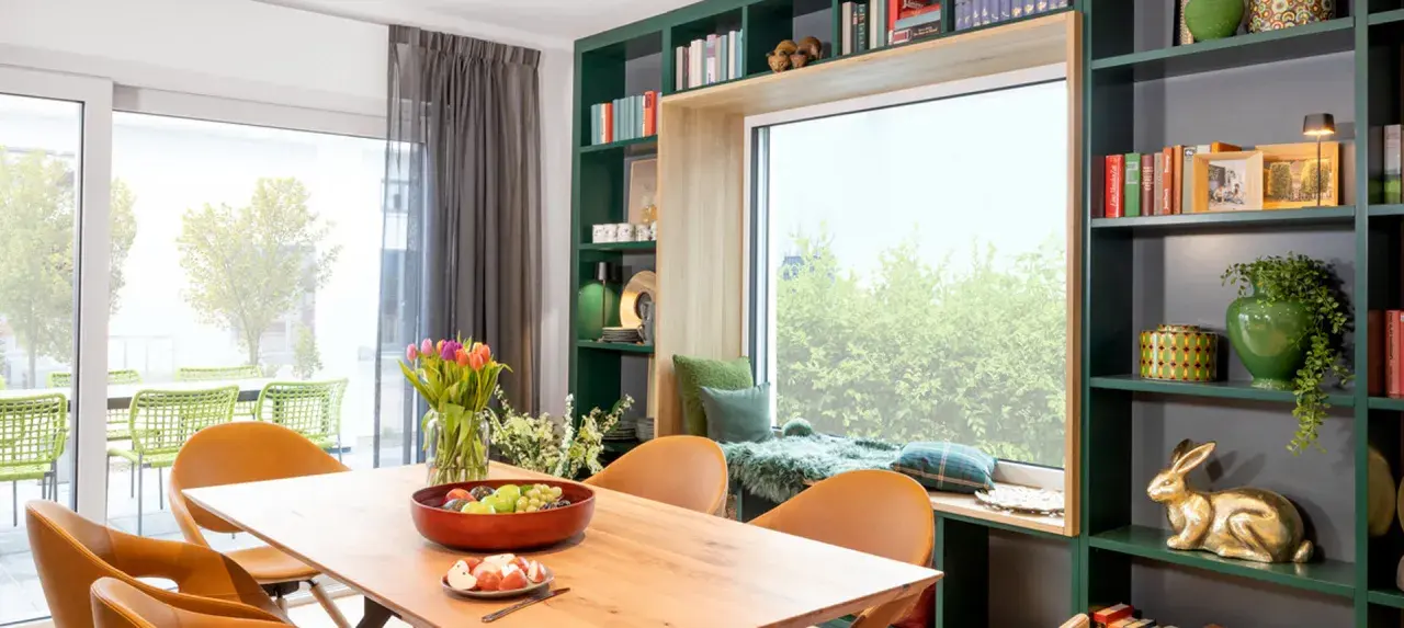 Gemütliche Leseecke mit Kissen und Decke am Fenster gestalten. Ein Holztisch mit Obstschale und Tulpen steht davor. Grüne Regale mit Büchern und Deko umrahmen das Fenster.