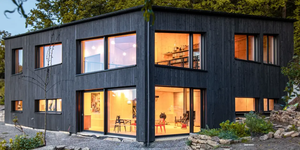 Modernes Haus mit Fassade im Shou Sugi Ban Stil. Große Fenster lassen warmes Licht herein.