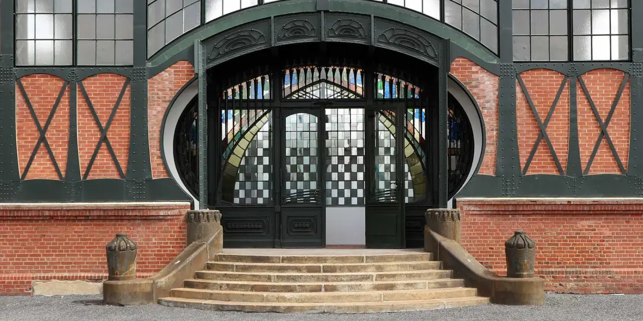 Eingang im artdeco stil mit Ziegelmauerwerk, geschwungenen Metallverzierungen und einem beeindruckenden Buntglasfenster.