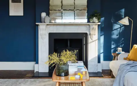 Ein Wohnzimmer mit tiefblauen Wänden. Ein Marmorkamin mit Spiegel darüber. Ein Holztisch mit Blumen und Spielzeug. Passt beige zu blau? Ja, die gelben Akzente und der beigefarbene Teppich zeigen es.