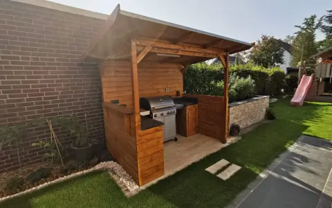 Ein moderner Grillplatz im Garten mit überdachter Holzkonstruktion, Gasgrill und gemütlicher Beleuchtung. Perfekt für Sommerabende.