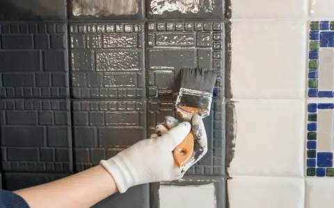Hand mit Handschuh streicht mit Pinsel graue Farbe auf Fliesen. Ein Teil der Wand ist bereits grau, der andere noch weiß.