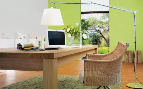 Ein Schreibtisch aus Nussbaumholz mit Laptop und Stoffmustern. Grüne Wand und Rattanstuhl. Welche Farbe passt zu Nussbaum? Grün und Beige.
