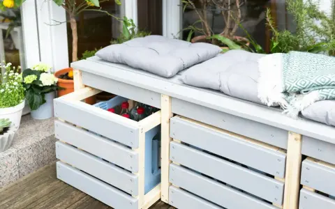 Ordnung im Garten: Eine graue Gartenbank mit Stauraum für Getränkekisten und Kissen. Pflanzen und eine Decke sorgen für Gemütlichkeit.