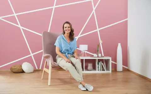 Frau sitzt vor einer rosa Wand mit weißem geometrischem Muster. Tolle Ideen zum Wand streichen und Muster abkleben!