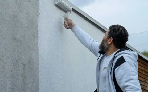 Ein Mann mit Bart streicht mit einer Malerrolle eine Hauswand. Die Wand zeigt horizontale Streifen, da sie gerade neu gestrichen wird.