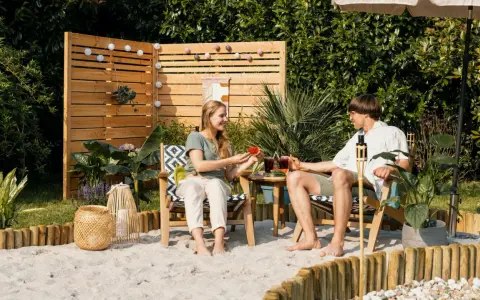 Ein Paar genießt den Sommer mit Wassermelone und Drinks im Garten. Sand, Holz und Pflanzen schaffen ein echtes Strandfeeling im Garten.
