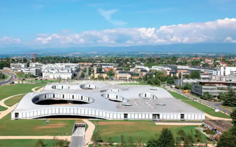 Das Rolex Learning Center in Lausanne, ein futuristisches Gebäude mit runden Öffnungen im Dach, umgeben von grünen Flächen und moderner Architektur.