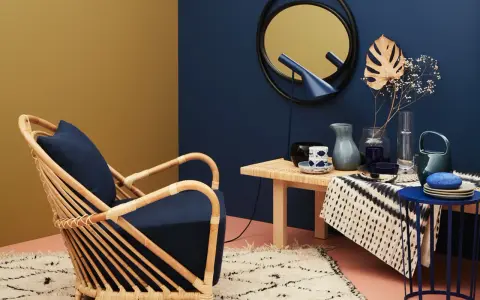 Ein Rattansessel mit dunkelblauem Kissen steht auf einem Teppich. Welche Farbe passt zu Gold? Das goldene Spiegelbild an der Wand und die gelbe Wandfarbe harmonieren mit dem tiefen Blau.
