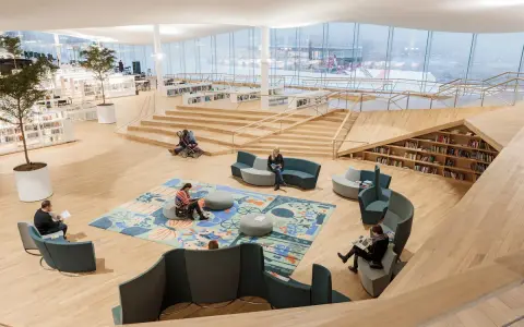 Helle, offene Bibliothek in Oodi Helsinki mit Menschen, die lesen und sich entspannen. Holzmöbel und große Fenster schaffen eine einladende Atmosphäre.