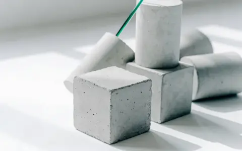 Betonwürfel und -zylinder in verschiedenen Formen, die die Vielfalt der **Beton festigkeitsklassen** symbolisieren. Ein grüner Stab ragt aus einem der Objekte.