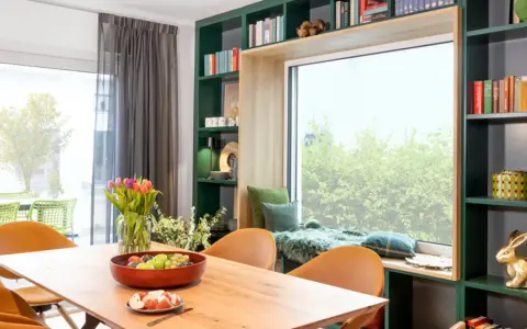 Gemütliche Leseecke mit Kissen und Decke am Fenster gestalten. Ein Holztisch mit Obstschale und Tulpen steht davor. Grüne Regale mit Büchern und Deko umrahmen das Fenster.