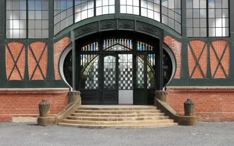 Eingang im artdeco stil mit Ziegelmauerwerk, geschwungenen Metallverzierungen und einem beeindruckenden Buntglasfenster.