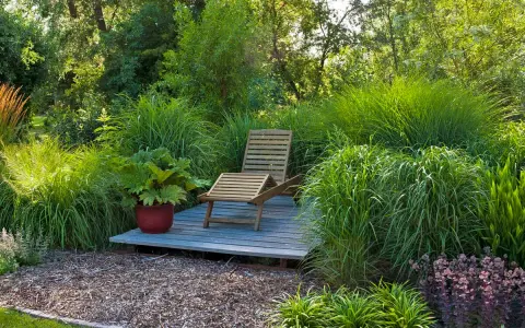 Entspannende Garten-Ideen: Eine Holzliege auf einer kleinen Terrasse inmitten üppiger Gräser und Pflanzen.