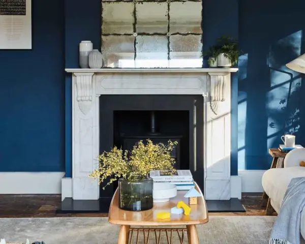 Ein Wohnzimmer mit tiefblauen Wänden. Ein Marmorkamin mit Spiegel darüber. Ein Holztisch mit Blumen und Spielzeug. Passt beige zu blau? Ja, die gelben Akzente und der beigefarbene Teppich zeigen es.