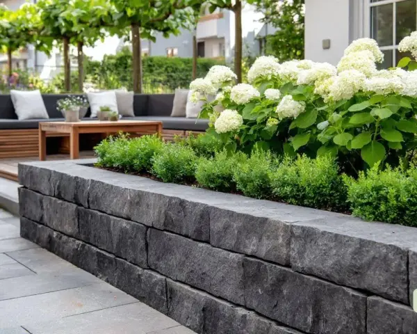 Moderne Terrasse mit Steinmauer und einem üppigen Beet. Ein gemütliches Lounge-Set lädt zum Entspannen ein.