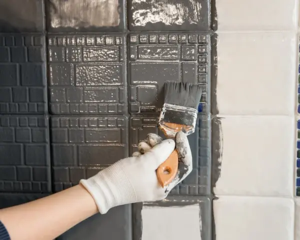 Hand mit Handschuh streicht mit Pinsel graue Farbe auf Fliesen. Ein Teil der Wand ist bereits grau, der andere noch weiß.