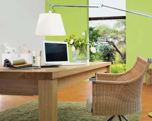 Ein Schreibtisch aus Nussbaumholz mit Laptop und Stoffmustern. Grüne Wand und Rattanstuhl. Welche Farbe passt zu Nussbaum? Grün und Beige.