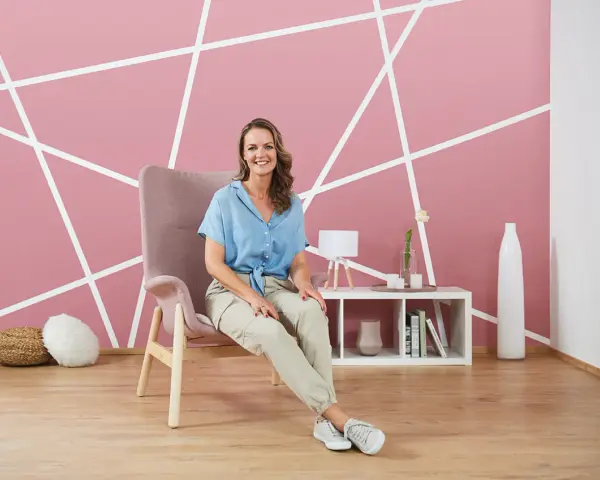 Frau sitzt vor einer rosa Wand mit weißem geometrischem Muster. Tolle Ideen zum Wand streichen und Muster abkleben!