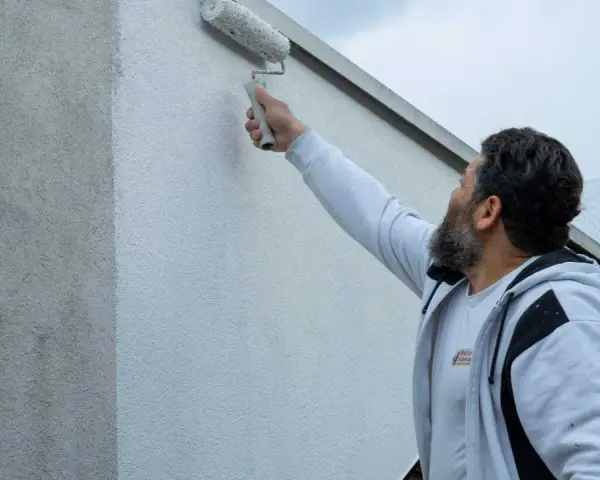 Ein Mann mit Bart streicht mit einer Malerrolle eine Hauswand. Die Wand zeigt horizontale Streifen, da sie gerade neu gestrichen wird.
