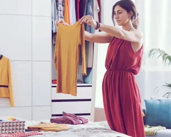 Frau sortiert Kleidung nach der Marie Kondo Methode. Ein gelbes Oberteil wird in die Hand genommen, daneben ein gelber Blazer und ein Korb mit gefalteten Textilien.