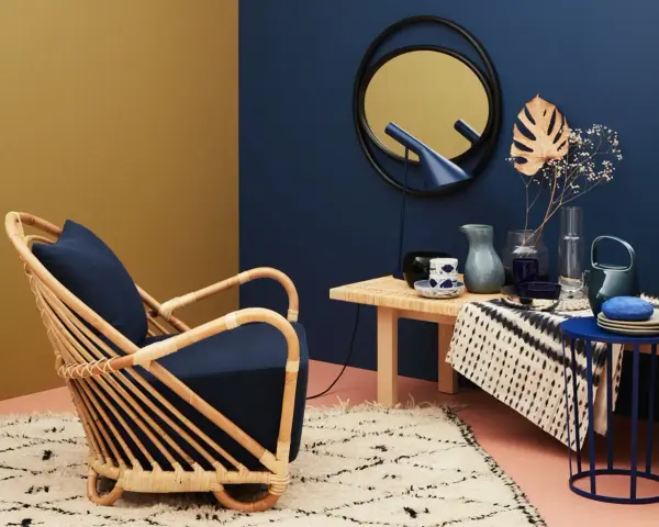 Ein Rattansessel mit dunkelblauem Kissen steht auf einem Teppich. Welche Farbe passt zu Gold? Das goldene Spiegelbild an der Wand und die gelbe Wandfarbe harmonieren mit dem tiefen Blau.