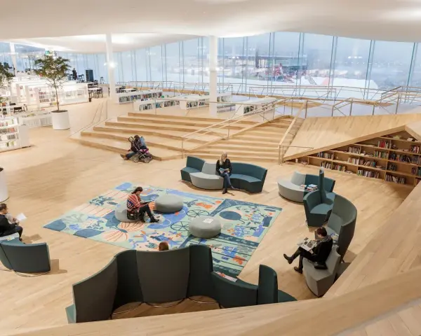 Helle, offene Bibliothek in Oodi Helsinki mit Menschen, die lesen und sich entspannen. Holzmöbel und große Fenster schaffen eine einladende Atmosphäre.