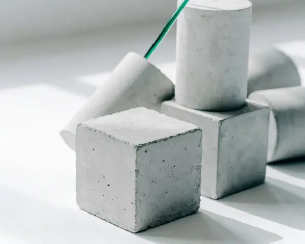 Betonwürfel und -zylinder in verschiedenen Formen, die die Vielfalt der **Beton festigkeitsklassen** symbolisieren. Ein grüner Stab ragt aus einem der Objekte.