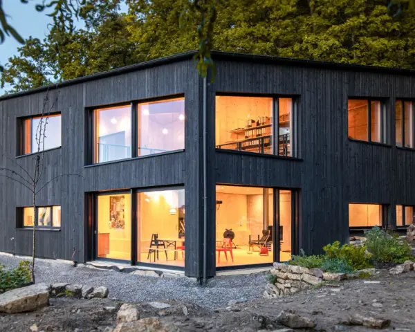 Modernes Haus mit Fassade im Shou Sugi Ban Stil. Große Fenster lassen warmes Licht herein.